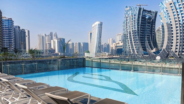 A rooftop pool with lounge chairs, a city skyline in the background, and modern buildings with the word "DUBAI" on one of them. - PVH - Exquisite 1BR - Binghatti Canal (Dubai)