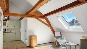 Una stanza in stile loft con travi a vista in legno, un velux, un angolo cottura, una zona salotto con tavolo e sedie, e un piccolo mobiletto.