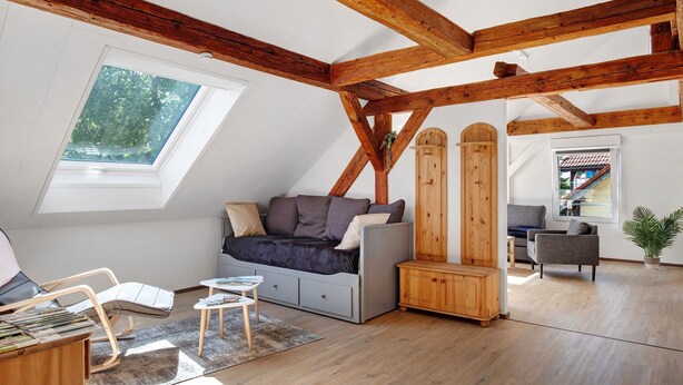 Una camera in stile loft con soffitto spiovente, travi in legno, un divano, una poltrona, un tavolino e una finestra vista alberi.