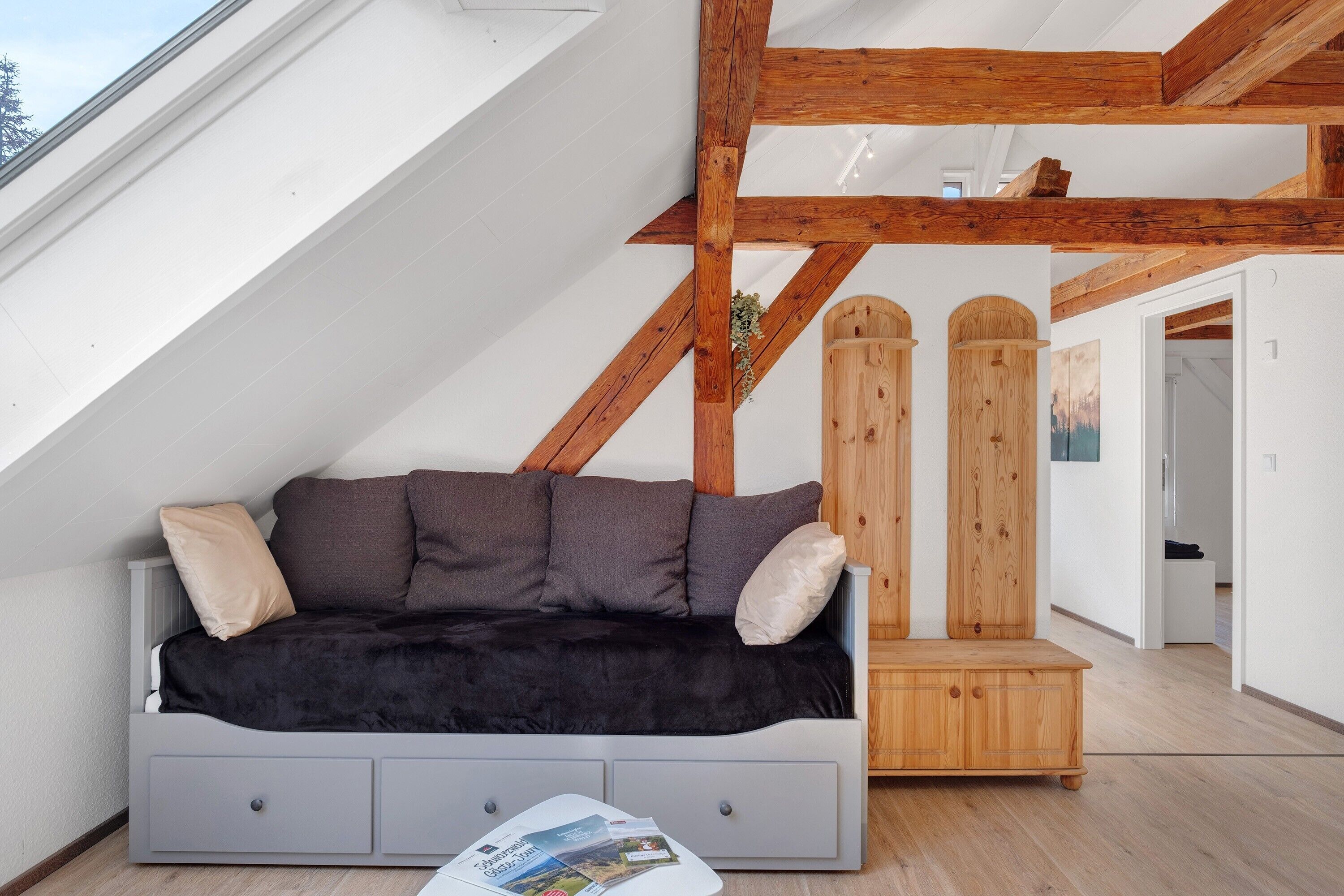 A loft-style room with a grey sofa, wooden beams, a small wooden cabinet, and a skylight.