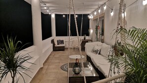 Un balcón con sofá blanco, una silla de mimbre, luces de macramé colgantes y plantas en macetas.