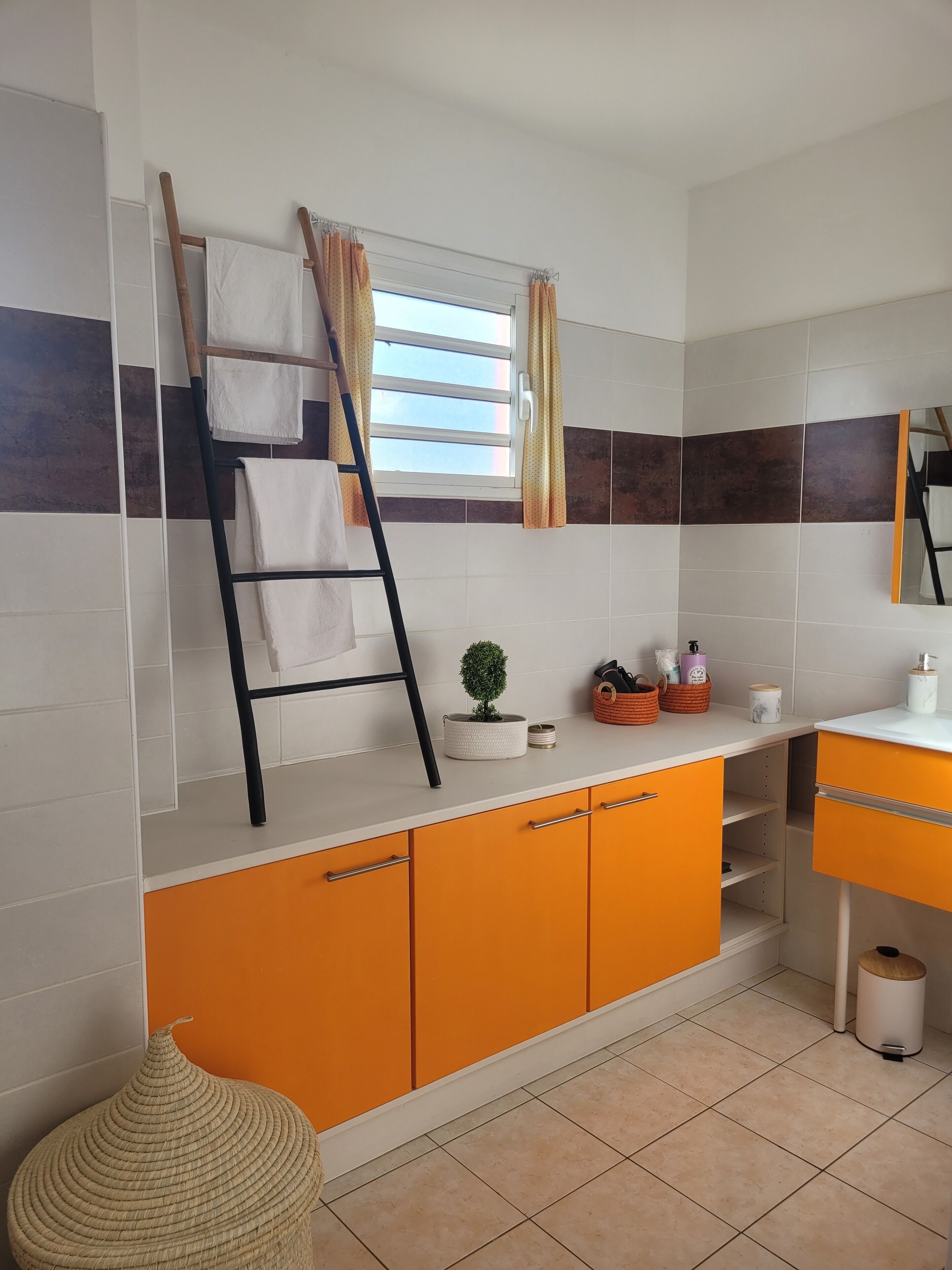 A bathroom with a tiled wall, a window with curtains, a towel rack, a small potted plant, and a sink with toiletries.