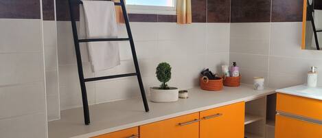 A bathroom with a tiled wall, a window with curtains, a towel rack, a small potted plant, and a sink with toiletries.
