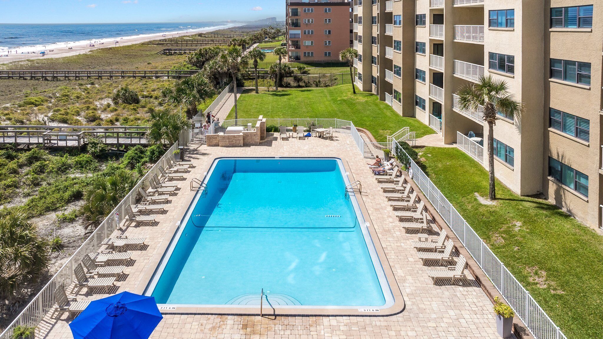 Une piscine rectangulaire à l’eau d’un bleu limpide, entourée de chaises longues et de parasols, avec une vue sur la plage et l’océan en arrière-plan.