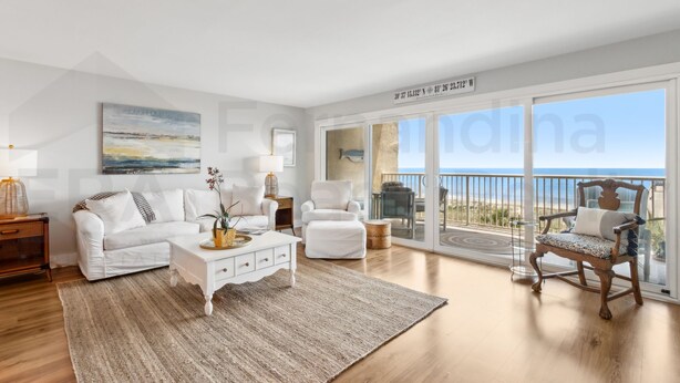A spacious living room with a white sofa, a coffee table, and a chair. There is a large window with a view of the ocean.