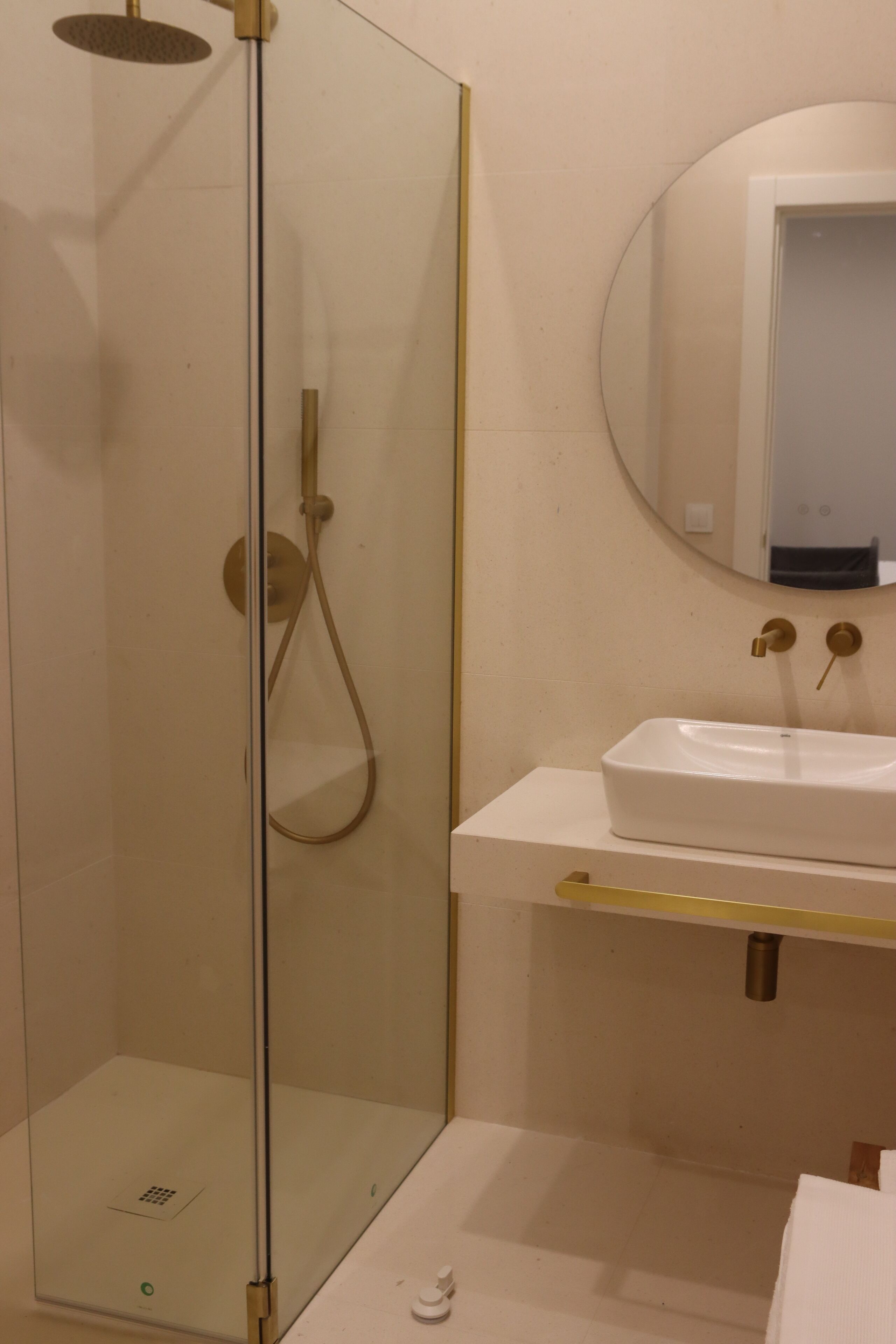 Un baño con ducha de vidrio, un lavamanos blanco con detalles dorados y un espejo redondo sobre él.