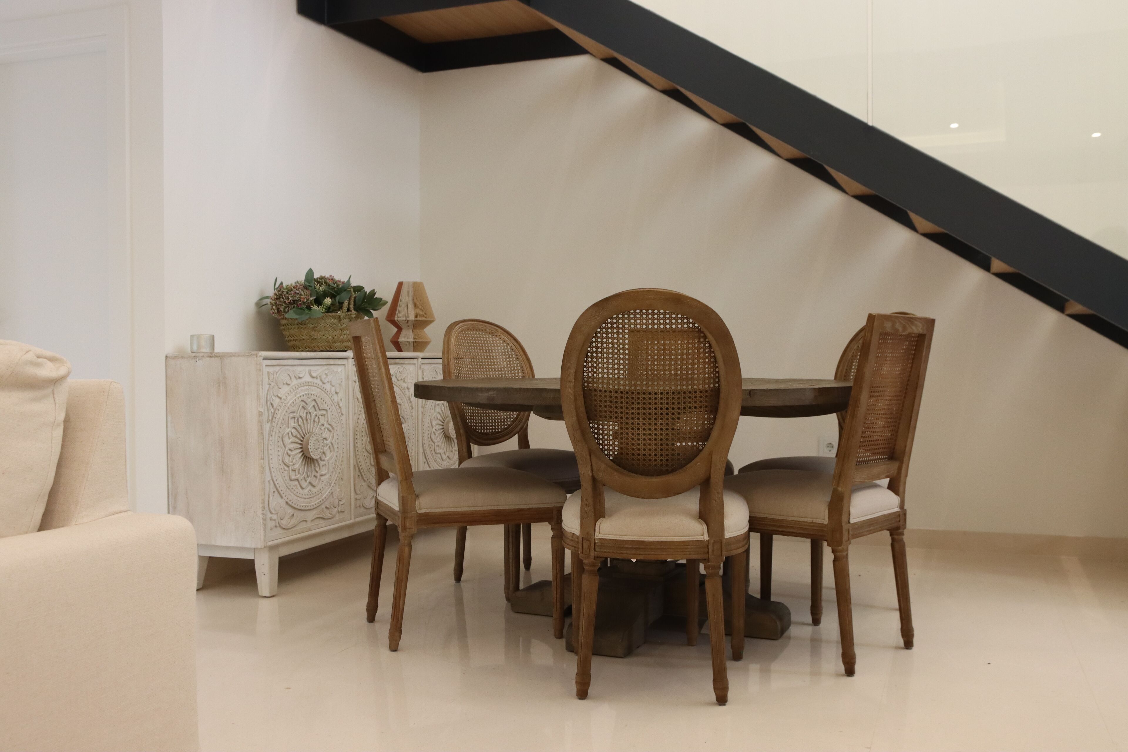 A dining area with a round table and four chairs, a white cabinet with decorative carvings, and a staircase in the background.