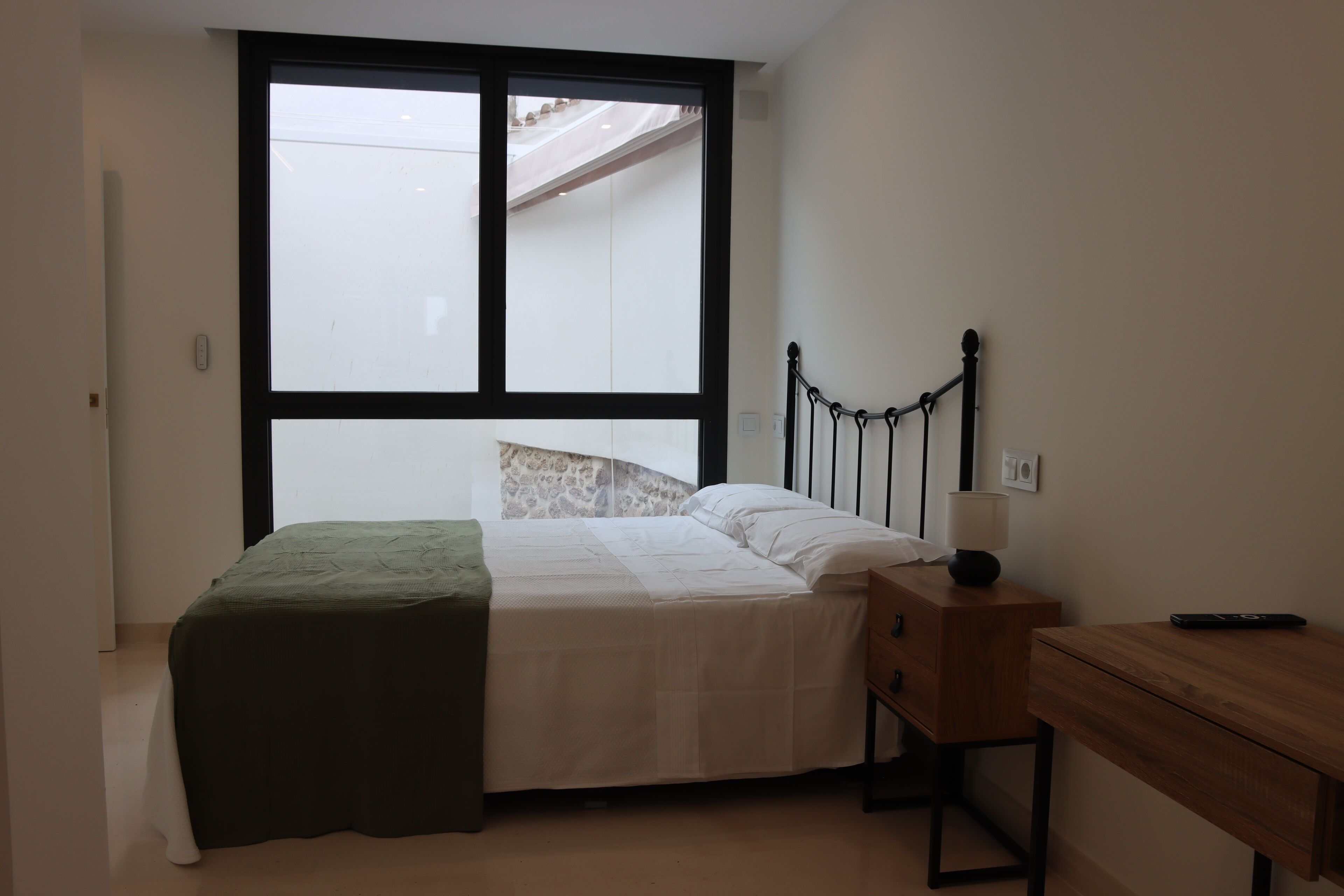 A bedroom with a bed, a nightstand, a desk, and a large window.