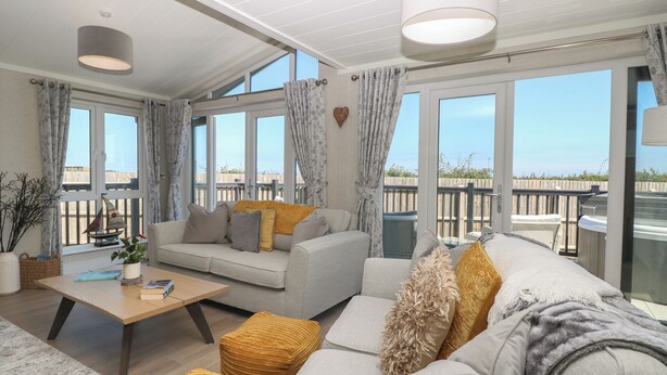 A spacious living room with a sofa, ottomans, and a coffee table. Large windows offer a view of the outdoors. - Life's a Beach (Norwich)