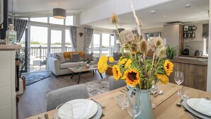 A modern living room with a sofa, coffee table, and a dining area set for two with a vase of sunflowers. - Life's a Beach (Norwich)