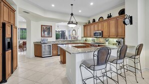 A spacious kitchen with granite countertops, wooden cabinets, and a large island with bar stools. - Castillo in Tiburon - Luxury Penthouse (Naples)