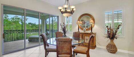 A dining area with a round glass table, four upholstered chairs, a chandelier, a wooden cabinet with a mirror, and a vase with dried plants.