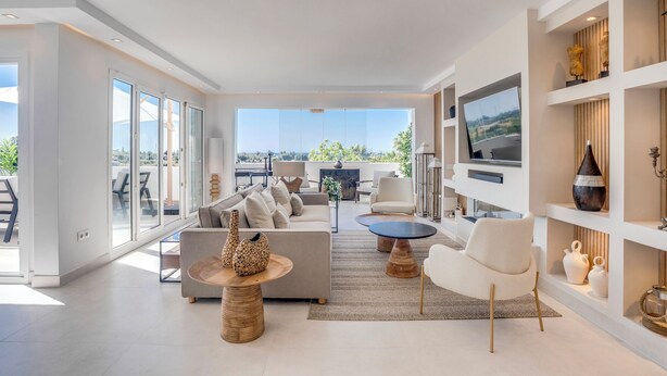 A spacious living room with a large sofa, a coffee table, and a TV mounted on the wall. There is a balcony with a view of the landscape.