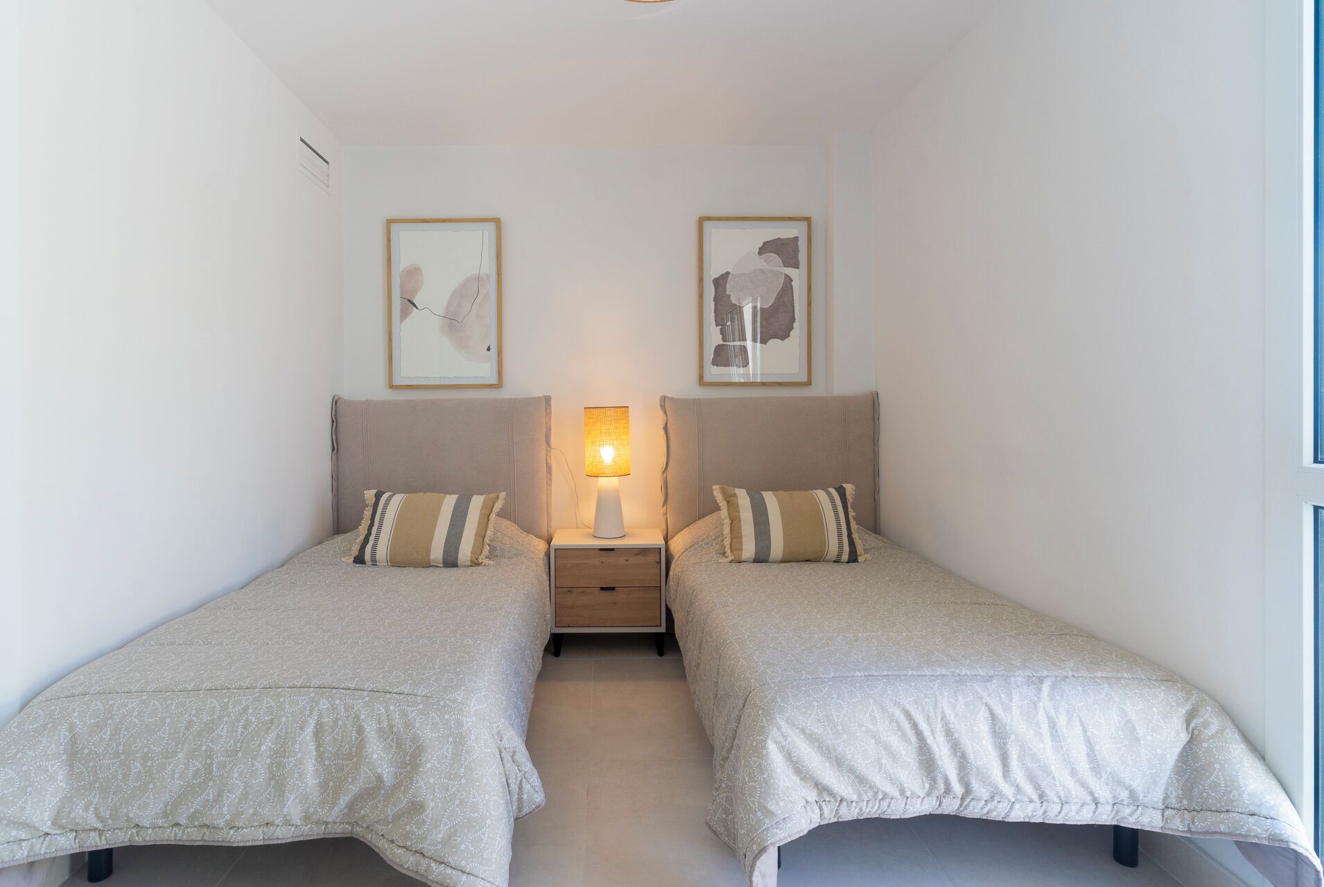 A bedroom with two single beds, each with a pillow and a bedside table with a lamp. There are two framed artworks on the wall above each bed.