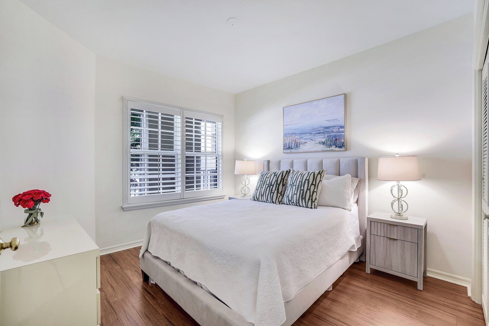 A bedroom with a large bed, white walls, a window with blinds, a bedside table with a lamp, and a painting above the bed.