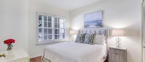 A bedroom with a large bed, white walls, a window with blinds, a bedside table with a lamp, and a painting above the bed.