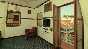 A room with a ceiling fan, a television mounted on the wall, a patterned floor, a window with a curtain, and a view of buildings outside. - 2BR in Stone Town – Near Ferry Terminal, Museums & Cultural Sights (Stone Town)