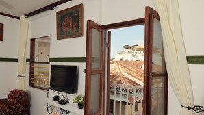 A room with a TV mounted on the wall, a window with a view of buildings, and a balcony with a railing. - 2BR in Stone Town – Near Ferry Terminal, Museums & Cultural Sights (Stone Town)