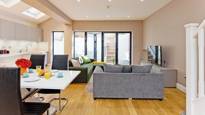A modern living room with a grey sofa, a dining table with breakfast items, and a kitchen area with cabinets and a TV. - Prime Central Base! 3BR/3BA Apt w/ Courtyard. Ideal for Families and Groups. (London)