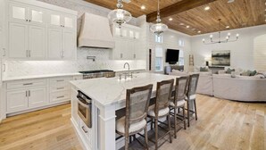 Une cuisine spacieuse avec des meubles blancs, un grand îlot central doté de tabourets, des poutres en bois au plafond et des luminaires suspendus.