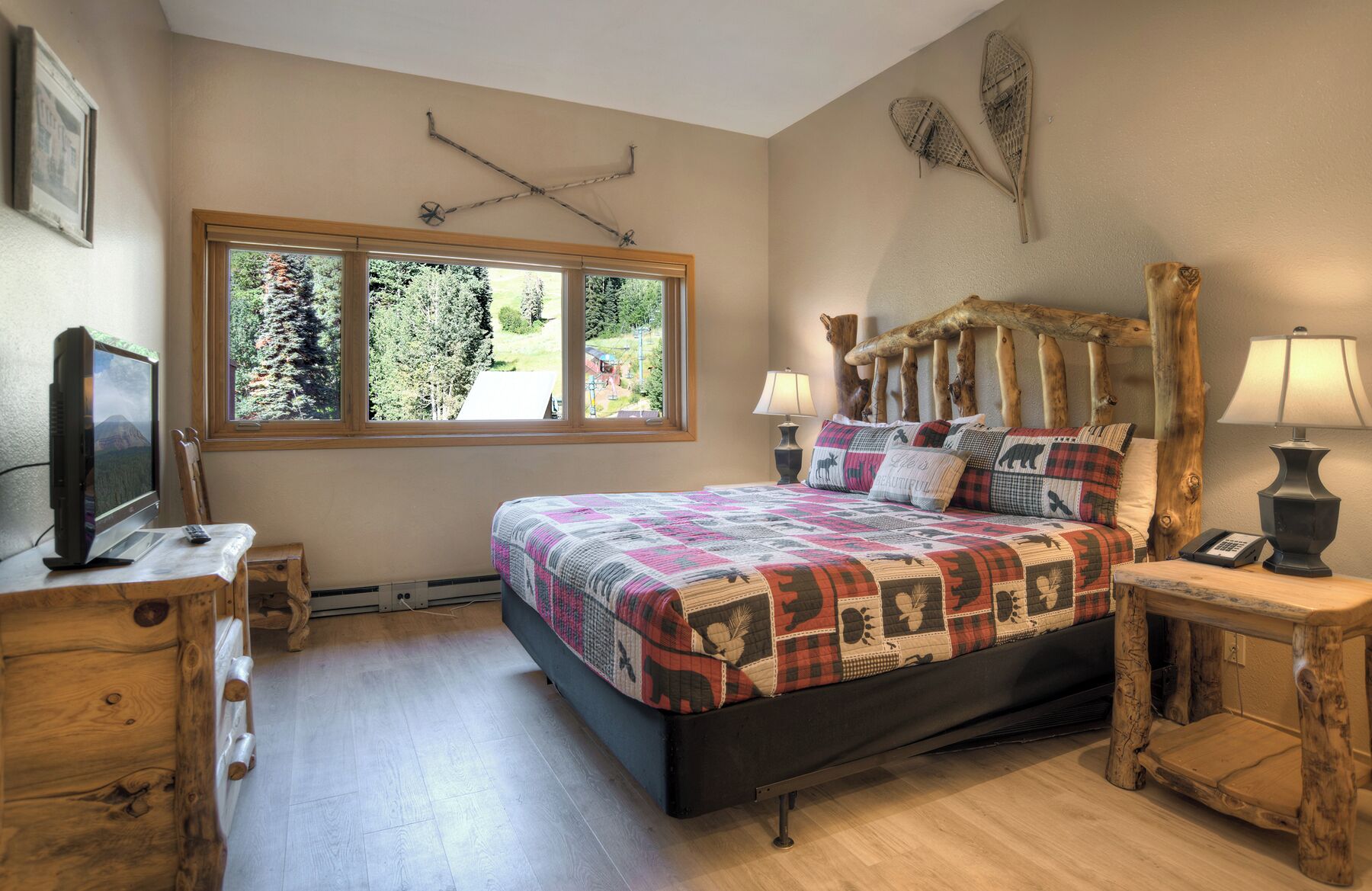 Ein Schlafzimmer mit großem Fenster, einem Bett mit Decke, einem Holz-Kopfende, einem Schreibtisch mit Lampe und einem Fernseher.