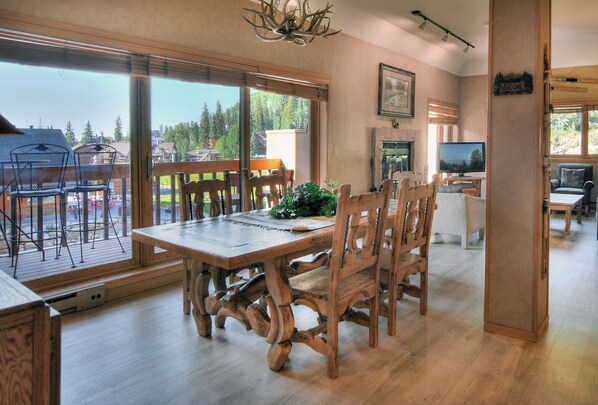A dining area with a wooden table and chairs, a television, and a view of a forest through large windows.