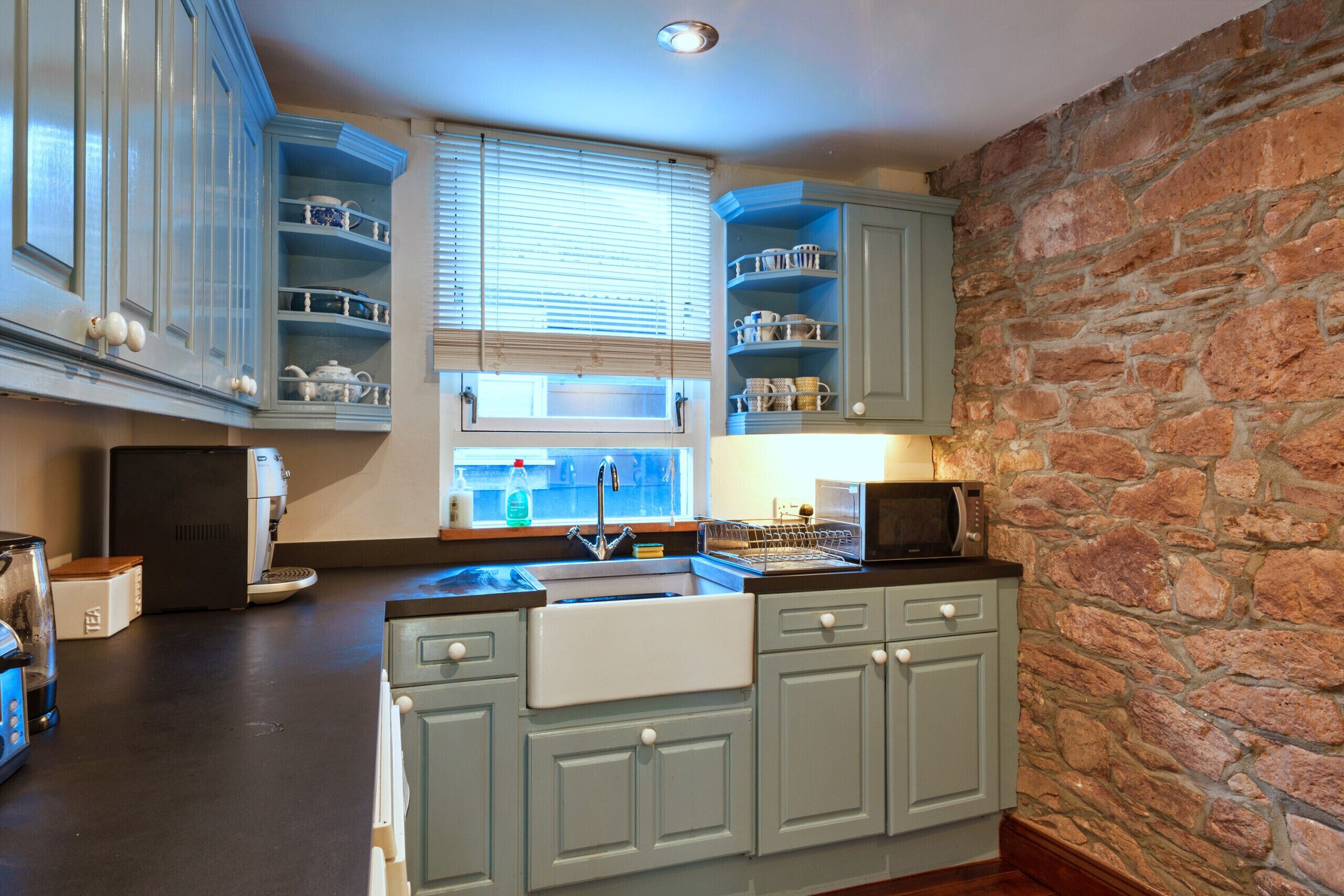 Une cuisine avec des meubles de couleur bleu clair, un mur en pierre, un four à micro-ondes encastré et un évier placé sous une fenêtre.