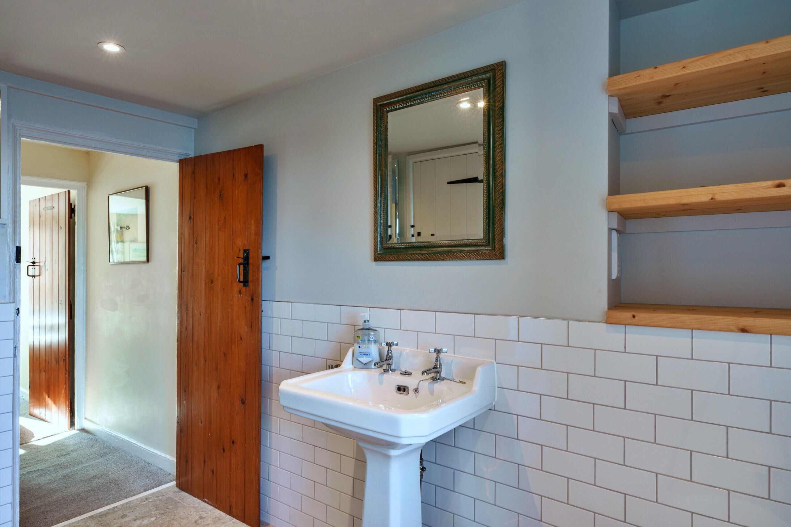 A bathroom with a white pedestal sink, a wooden door, and a mirror with a decorative frame.