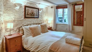 A bedroom with a stone wall, a bed with white bedding, two bedside tables with lamps, and a window with curtains. - Penlee Narrows, Kingsand (Kingsand)