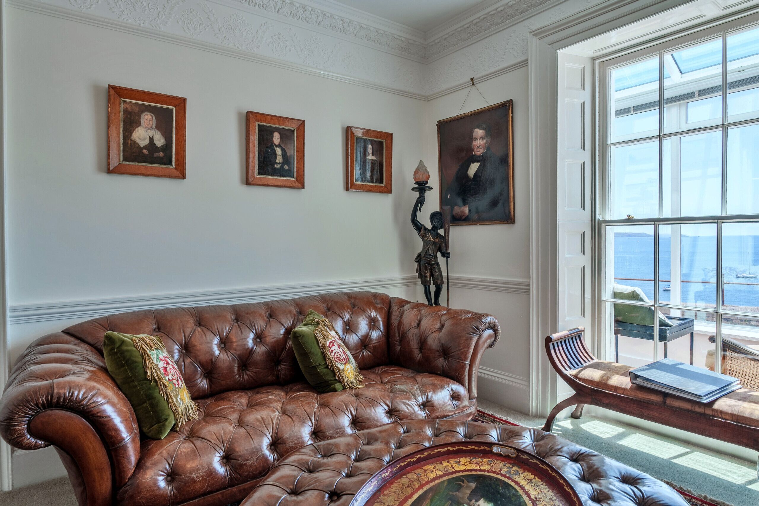 A room with a large brown leather sofa, a wooden side table, and three framed portraits on the wall.