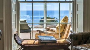 A room with a view of the ocean through a large window, a leather sofa, a wooden chair, and a small table with a book. - Penley Villa (Kingsand)