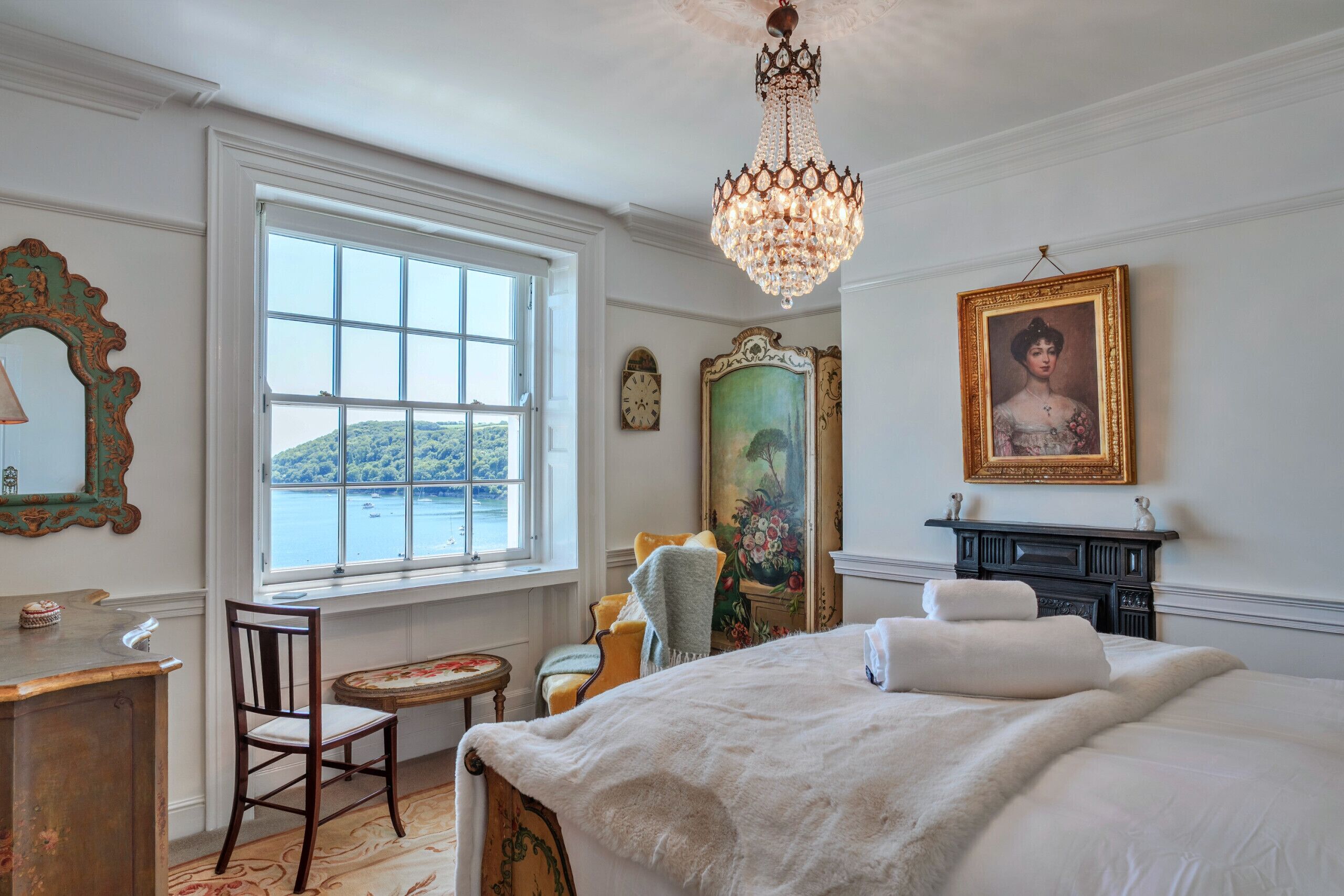 A bedroom with a large bed, a chandelier, a window with a view, a dresser, and a chair.