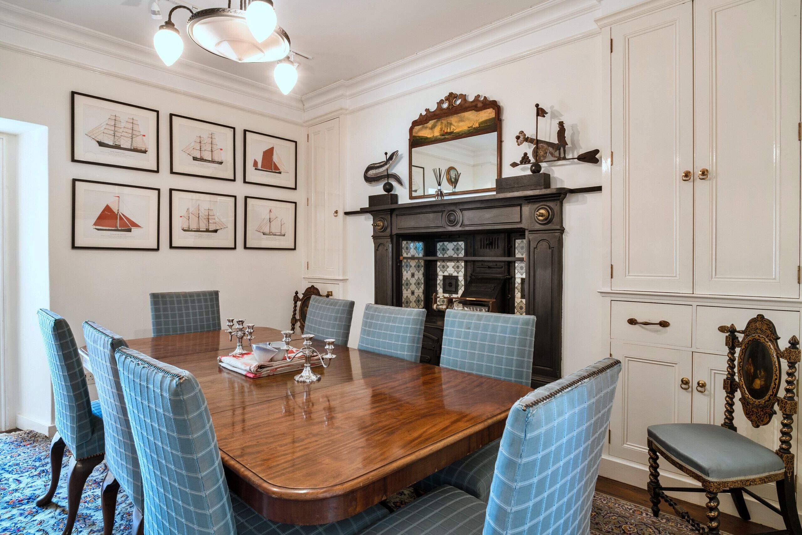 A dining room with a large wooden table surrounded by blue upholstered chairs, framed maritime artwork on the wall, and a fireplace with decorative items.