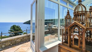 A room with a large glass window overlooking a coastal view, a wooden decorative piece, and a stone wall. - Penley Villa (Kingsand)