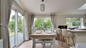 Une cuisine moderne avec une table en bois et des tabourets de bar, de grandes fenêtres avec des rideaux et une vue sur la verdure à l’extérieur.