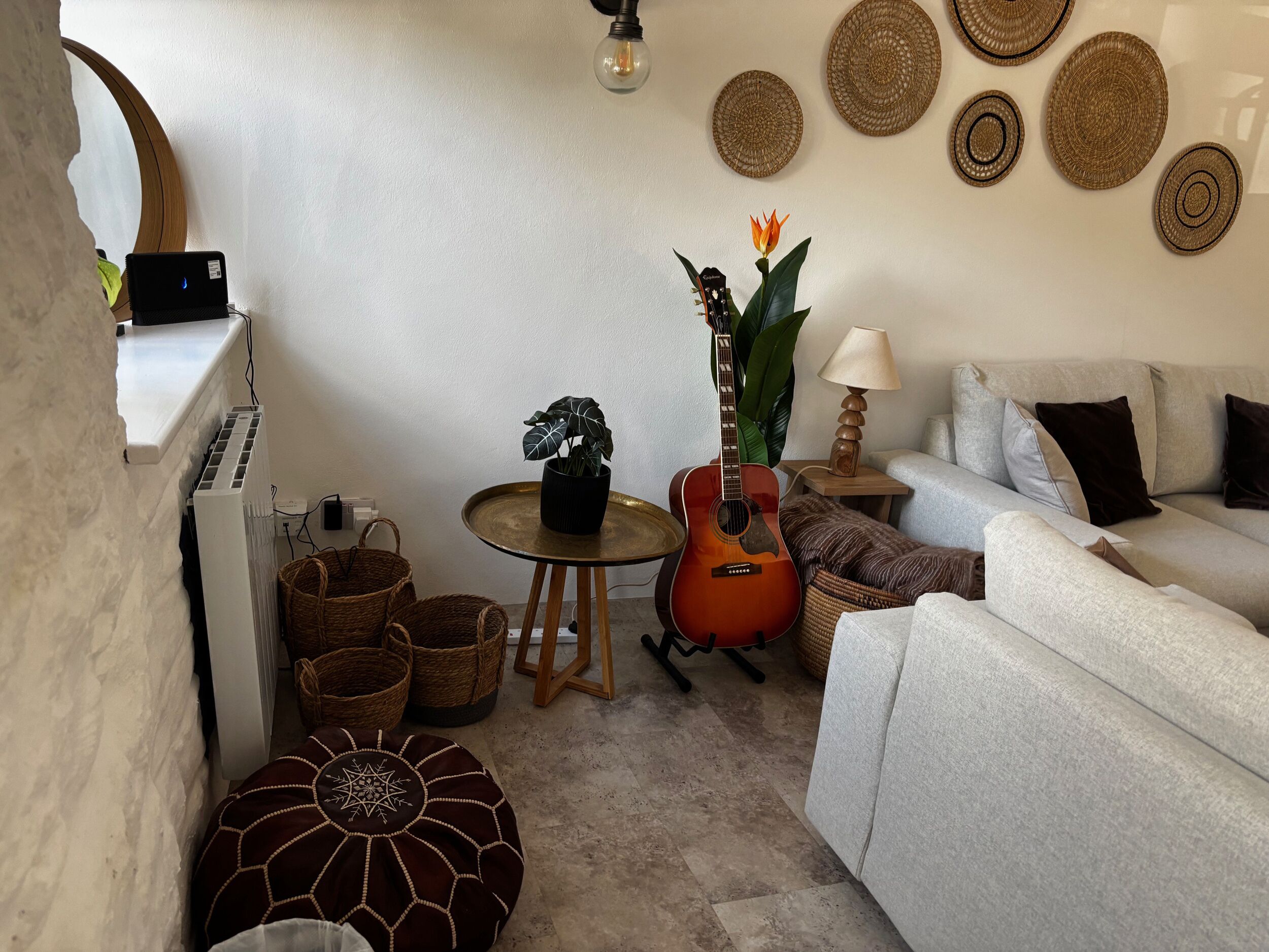 Una sala de estar acogedora con un sofá gris, una mesita redonda con una lámpara, una guitarra y una planta en maceta. En el suelo, se observan tapices y cestos de mimbre.