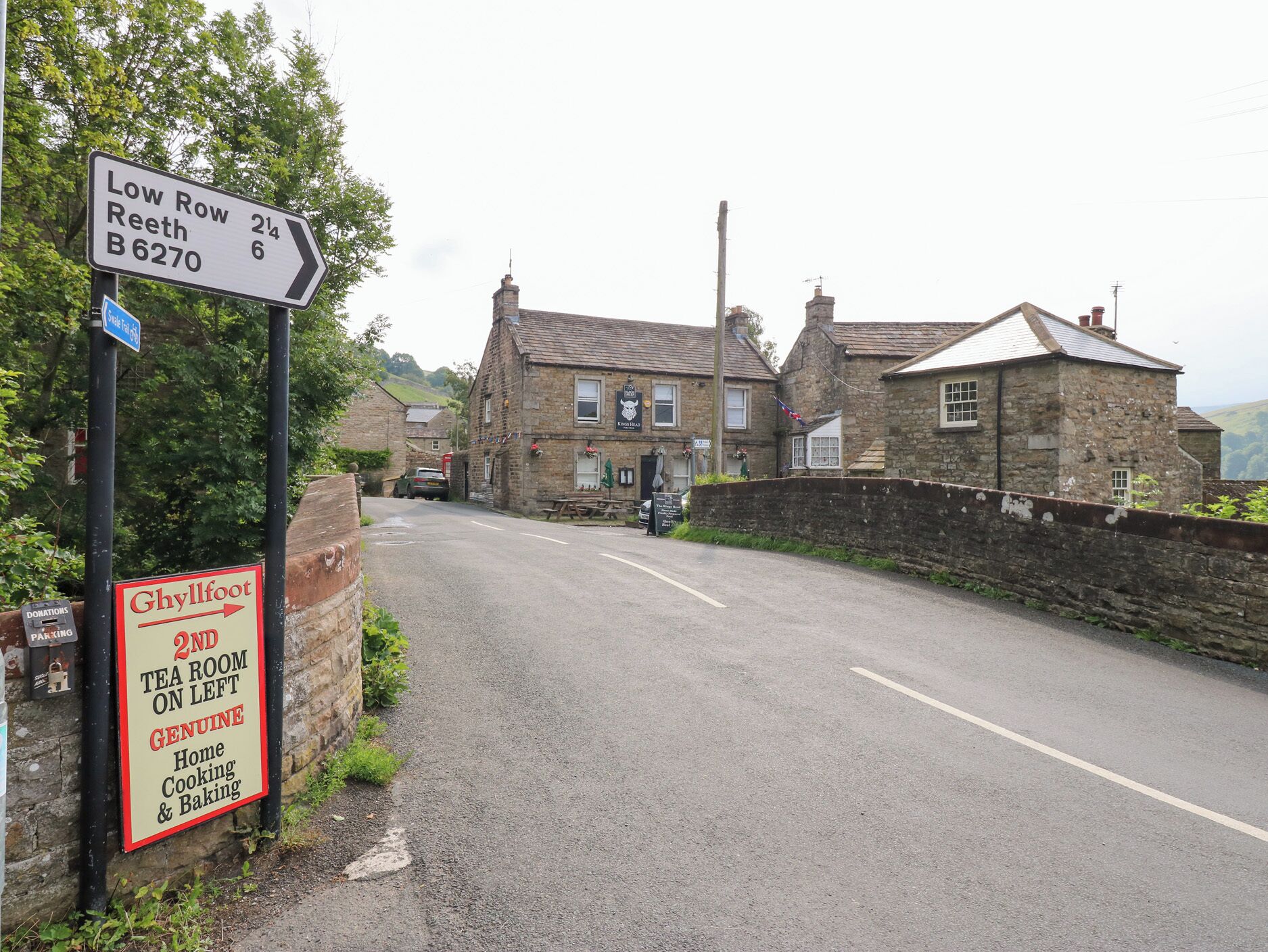 Une route de campagne avec un panneau indiquant « Low Row Reeth B6270 », un salon de thé sur la gauche et un pub appelé « King Head ».