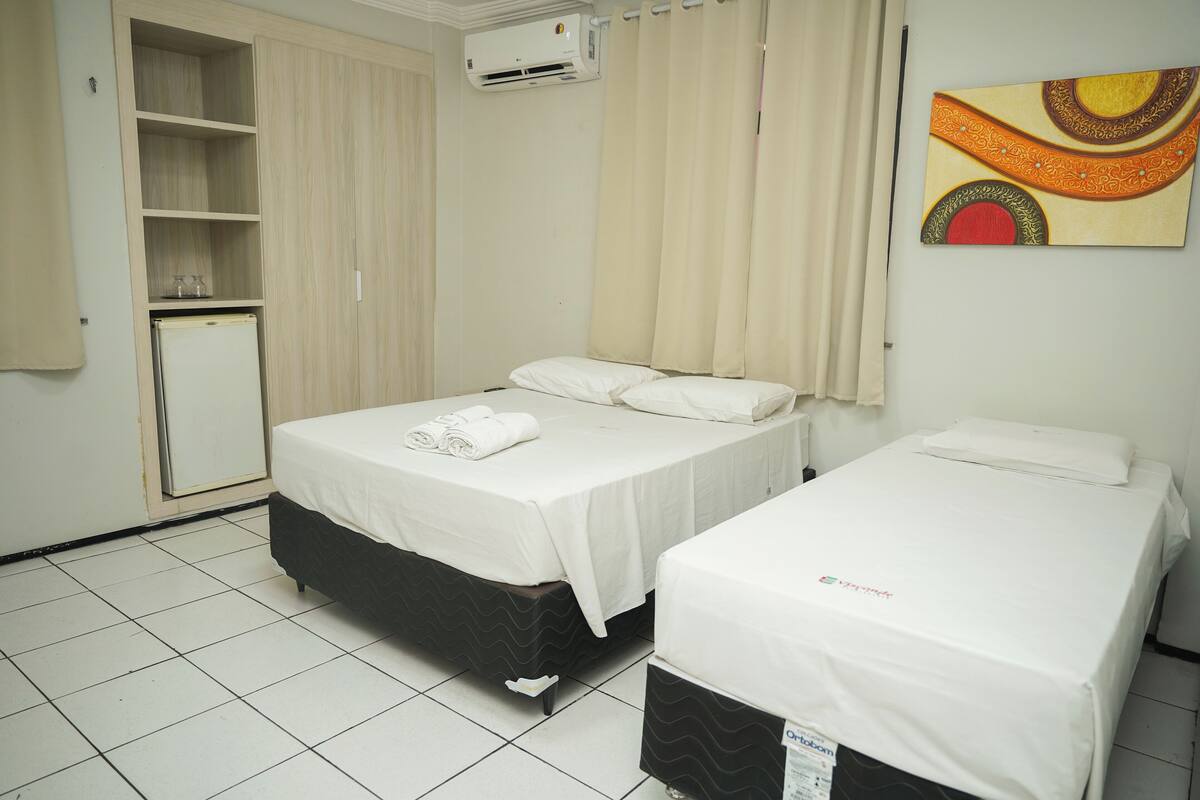A hotel room with two beds, a wardrobe, a small refrigerator, and an air conditioning unit.