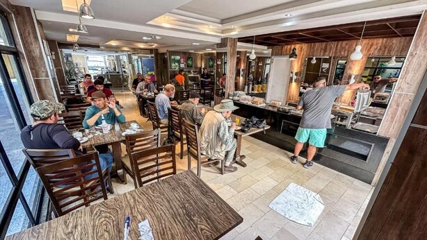 A restaurant with wooden tables and chairs, a counter with food items, and a person serving. - Hotel Visconde  (Sobral)