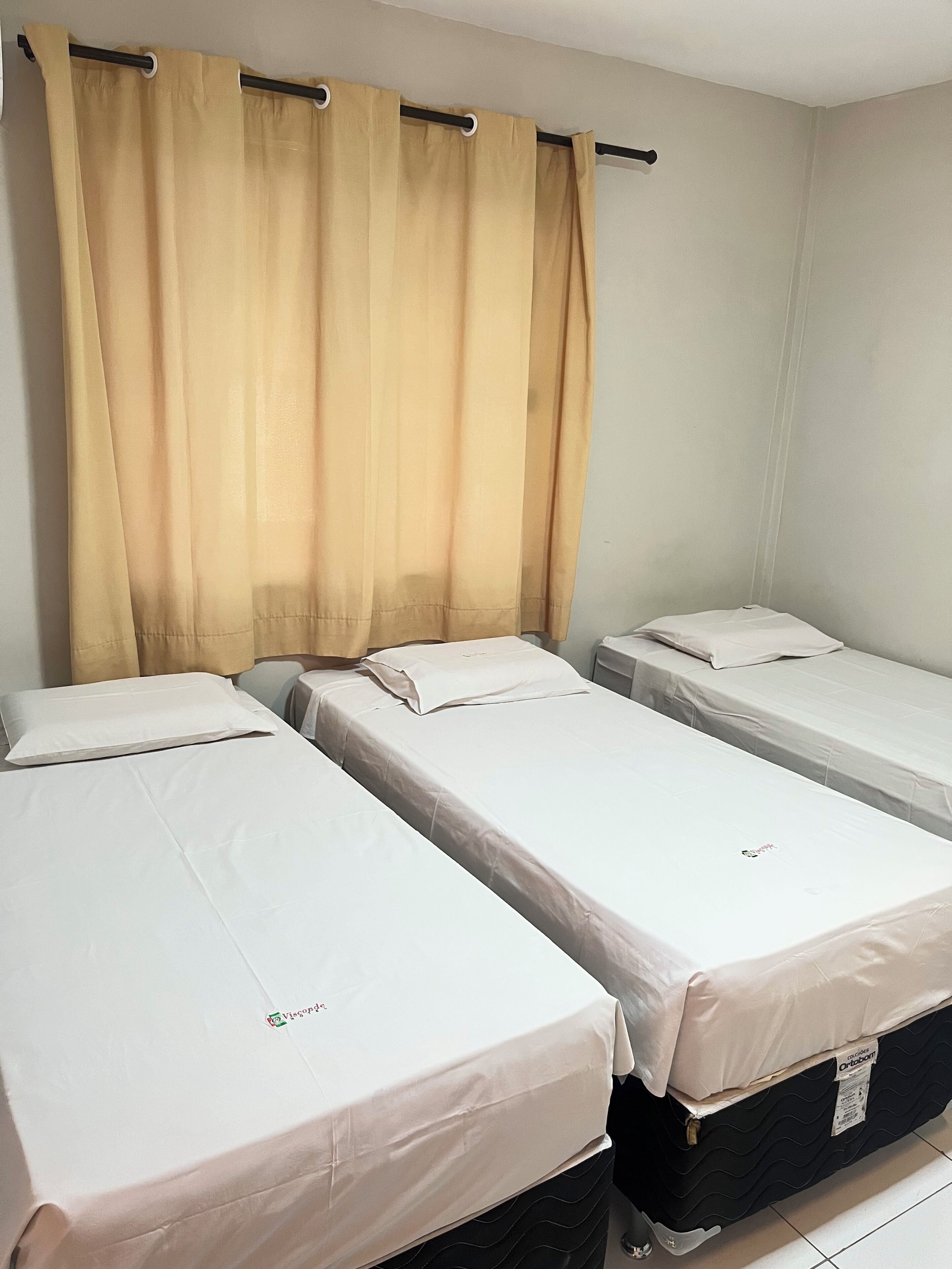 A room with three single beds, each with a white mattress and pillow. There is a beige curtain on a rod above the beds.