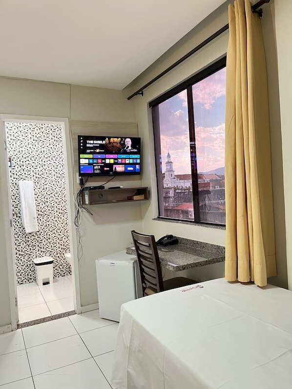 A hotel room with a bed, a small table, a TV mounted on the wall, and a window with curtains.
