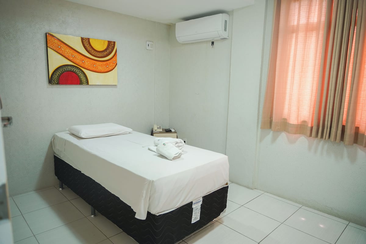 A hotel room with a bed, a wall-mounted air conditioner, a bedside table with a phone, and a window with curtains.