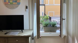 A room with a TV on a wooden desk, a potted plant by the window, and a view of a yellow building outside. - Aro's House (Cremona)