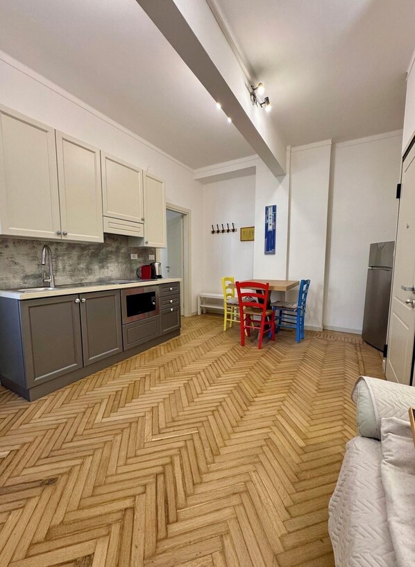 A kitchen with white cabinets, a herringbone-patterned floor, and a dining area with a red and blue chair. - Aro's House (Cremona)