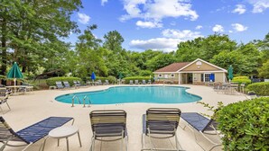 A pool area with lounge chairs and umbrellas, surrounded by trees and a building with a red roof. - 223 Greenbriar - Barefoot Resort Rentals (North Myrtle Beach)