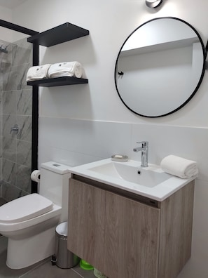 Une salle de bain moderne avec des toilettes, un lavabo et un miroir. Il y a des étagères au-dessus du lavabo et une poubelle en dessous.