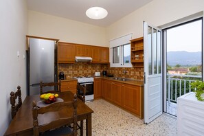 A kitchen with wooden cabinets, a stainless steel refrigerator, a dining table with chairs, and a view through a sliding glass door. - Valira Stathmos Home - Rustic Charm Cottage W Garden (Messini)