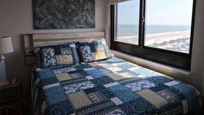 A bedroom with a large window overlooking a beach, a bed with a blue and beige quilt, and a painting on the wall. - Awesome II_Beach Views From the Bed (Myrtle Beach)