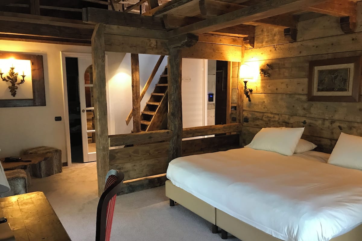 A bedroom with a wooden ceiling, a bed with white bedding, a chair, a wooden table, and a staircase leading to another room.