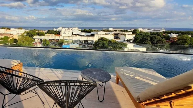 A rooftop pool area with lounge chairs and a small table, overlooking a cityscape with buildings and greenery. - Magnificent location, Rooftop with Ocean View (Playa del Carmen)